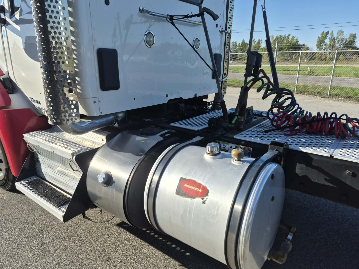 Kenworth T880 cab with fuel tank and steps; circular aluminum fuel tank with black band and two bolts; dashboard controls; red coiled lines; perforated metal side step and white cab; windshield wipers visible.