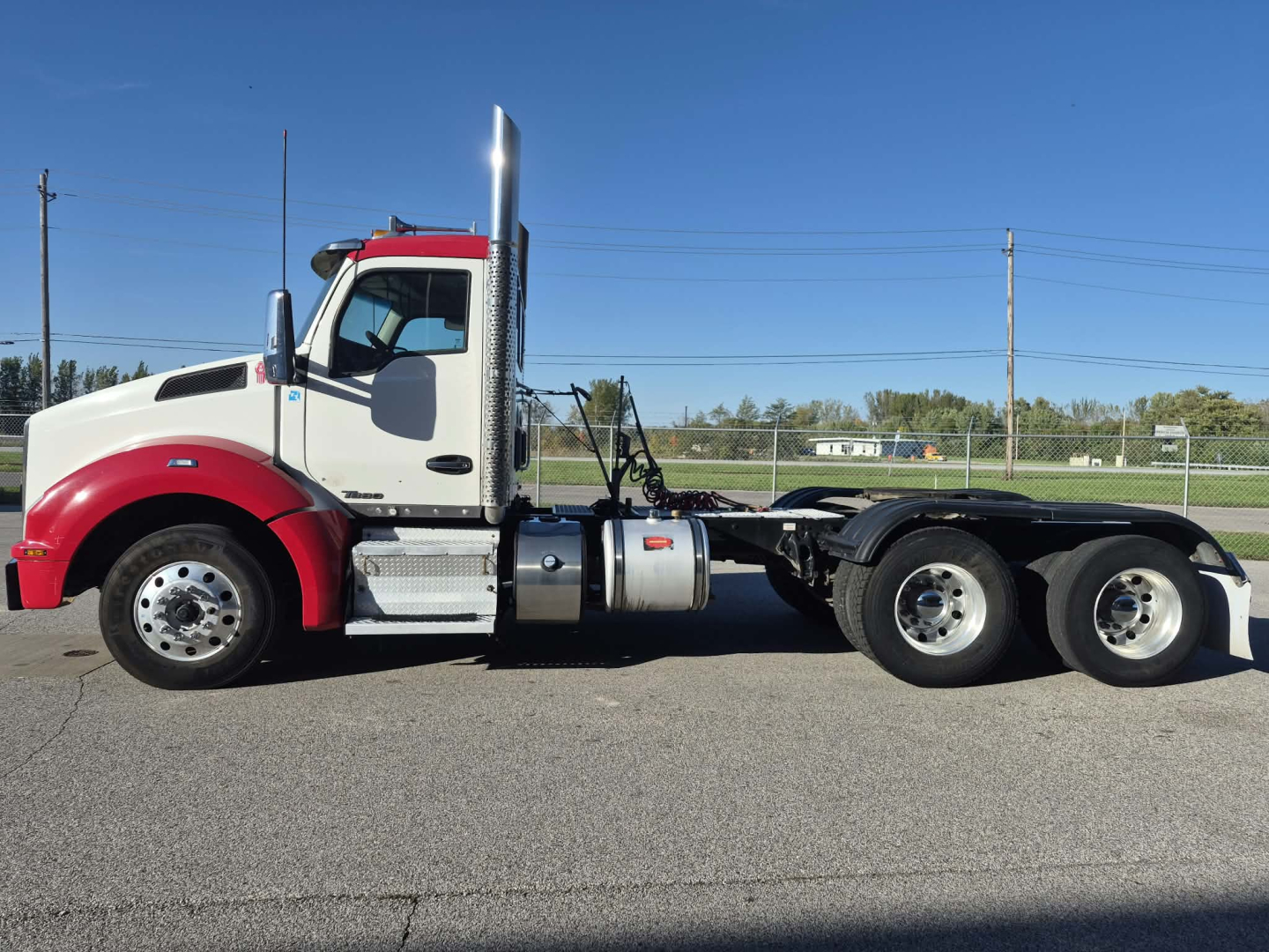 Kenworth T880 conventional daycab truck, side view showing red/white cab, chrome exhaust stack, dual rear axles, fuel tanks, and bare frame.