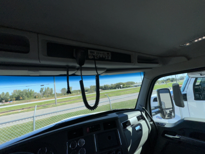 Kenworth T880 cab interior with overhead dashboard and CB microphone; windshield and side mirror visible; exterior road and fence in background.