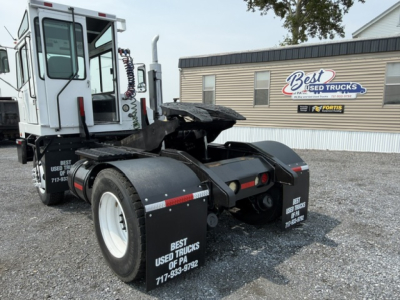 Truck tractor rear view showing fifth wheel hook-up and dual rear tires with fenders, dealership signage in background.