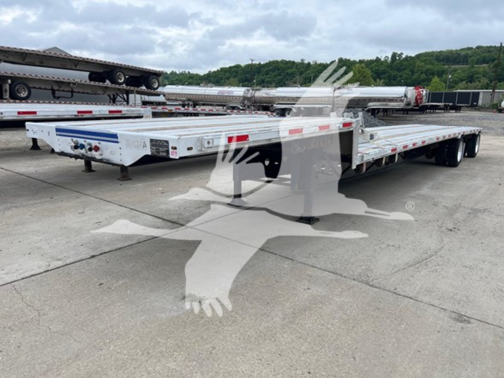Flatbed trailer with white deck and dual rear axles, low-profile hitch, and chrome wheels. Visible are side rails and reflective tape; background shows other trailers in a lot.