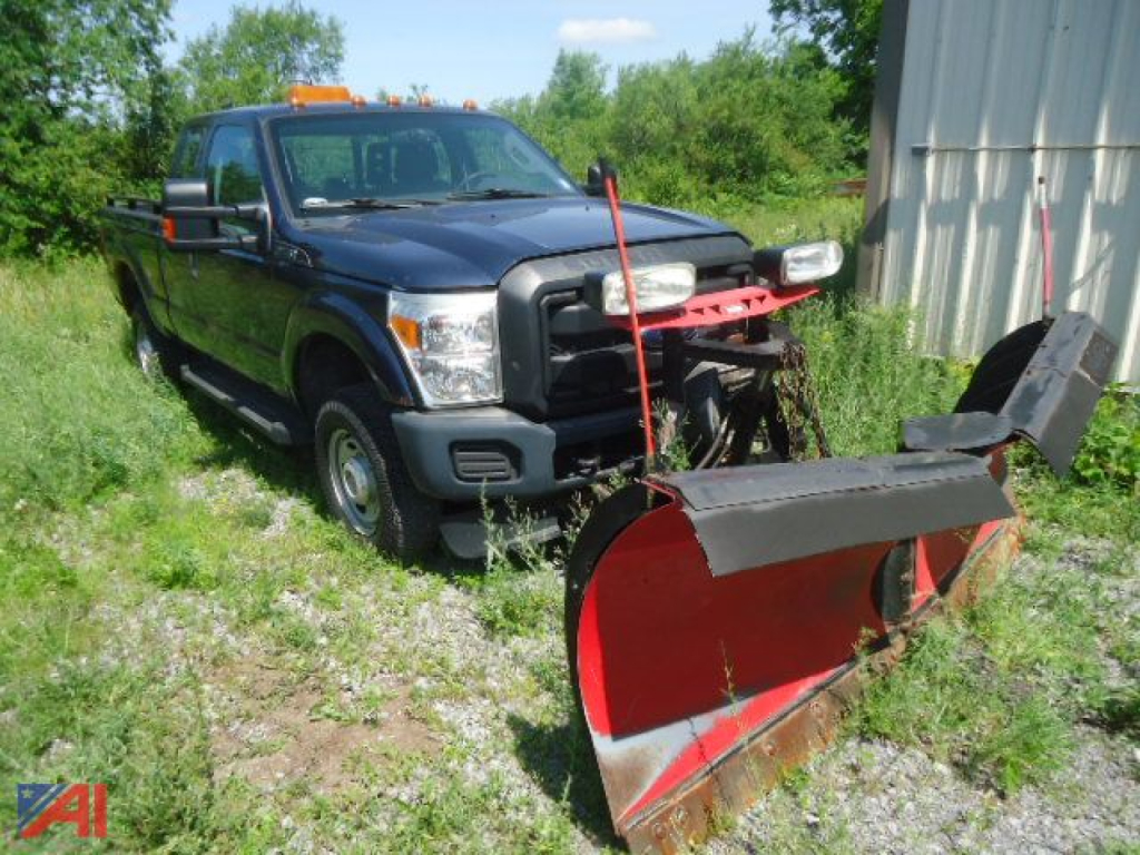 Ford F-250 Super Duty pickup with a red plow attachment in a field, blue truck, daytime, warning lights on roof, front grille visible.