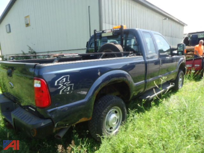 Ford F-250 Super Duty pickup with 4x4 V-Plow, blue extended cab, rear view showing tailgate and spare tire, parked beside a warehouse.