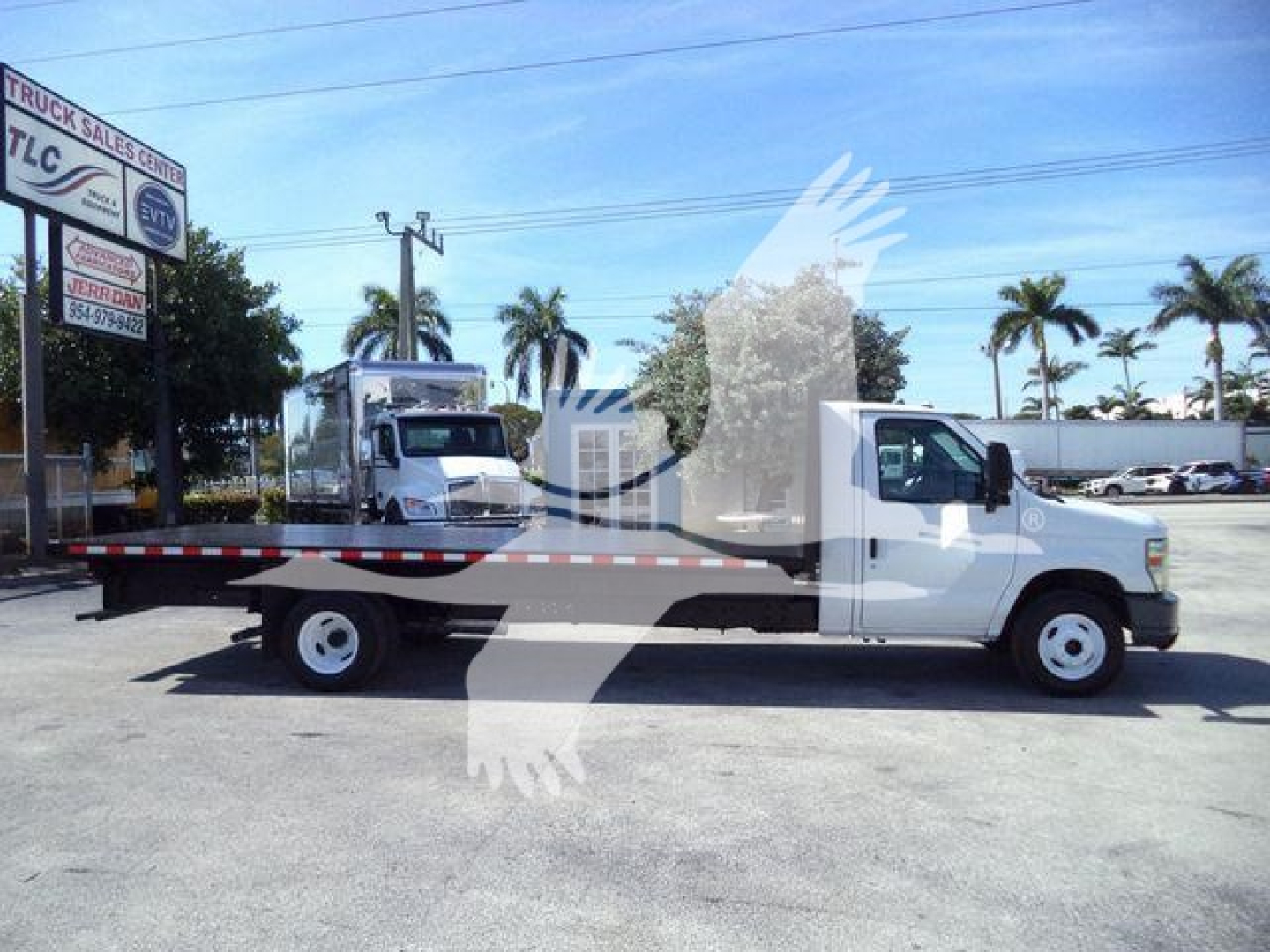 Flatbed tow truck side view showing long flatbed and white cab with branding on the door, palm trees in background.