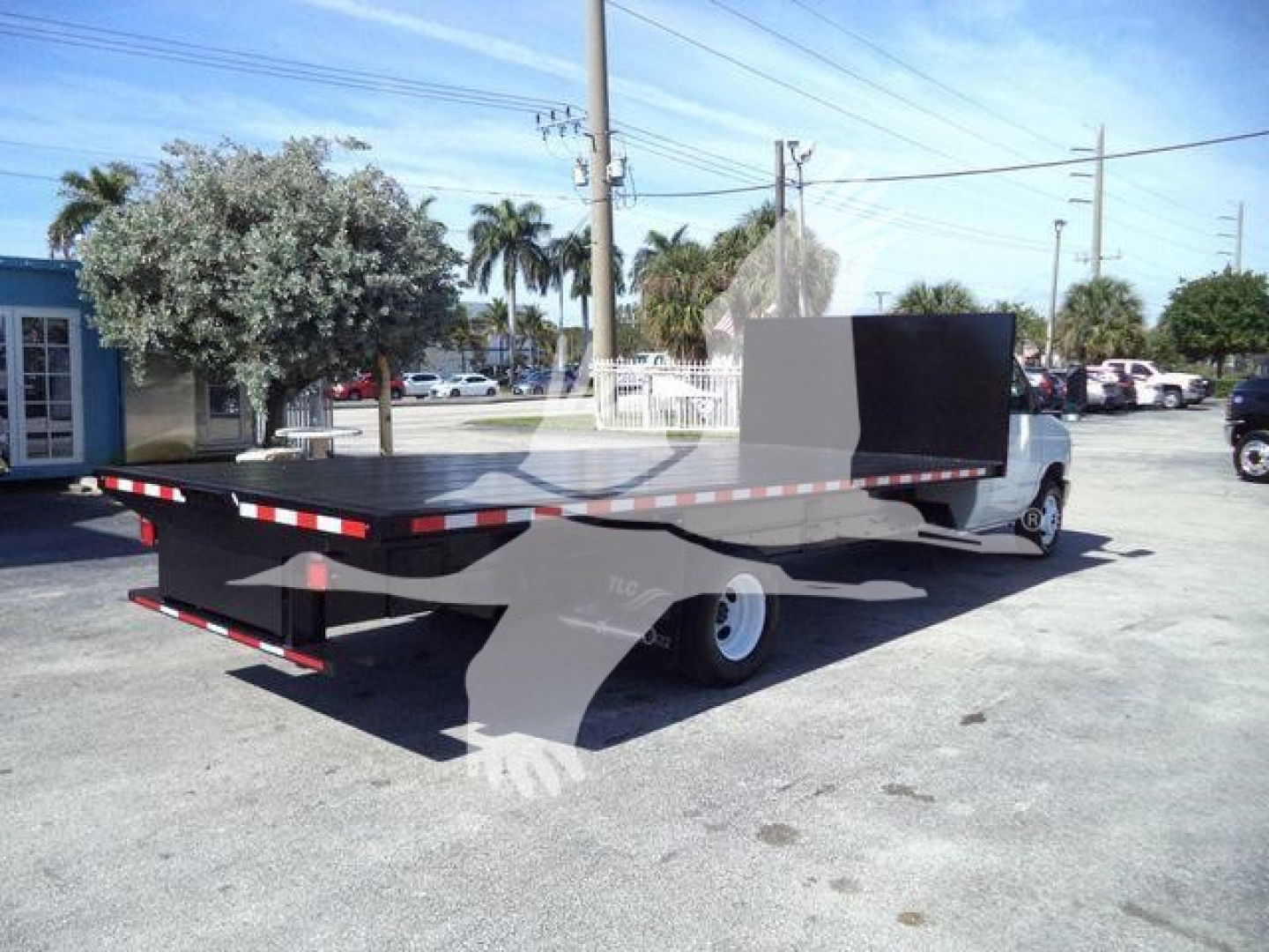 Flatbed tow truck on lot with long deck and rear drop, rear bumper, and white cab. Flatbed extends toward camera with black metal sides and a tall black barrier at cab end.