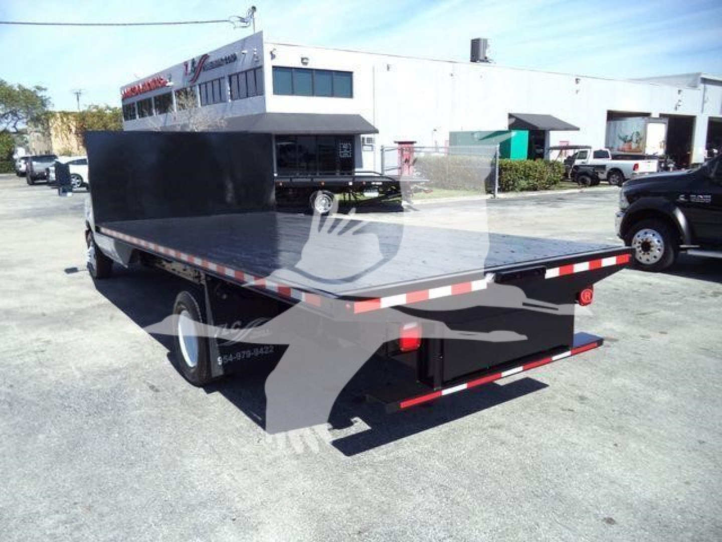 Flatbed tow truck bed with black coating and reflective tape on rear edge, viewed from rear-left side; stock number and dealership in background.