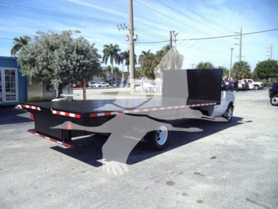 Flatbed tow truck on lot with long deck and rear drop, rear bumper, and white cab. Flatbed extends toward camera with black metal sides and a tall black barrier at cab end.