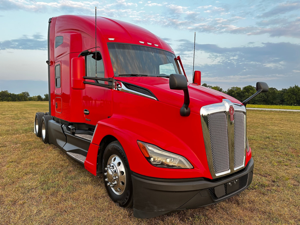 Kenworth T680 sleeper cab truck in red, front-left view with chrome grille and prominent front bumper, on grassy lot.