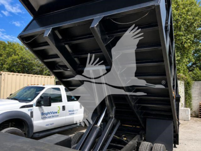 Isuzu NPR-HD dump truck bed raised in a yard with a white service truck in background; view focuses on hydraulic dumping mechanism and steel dump bed.