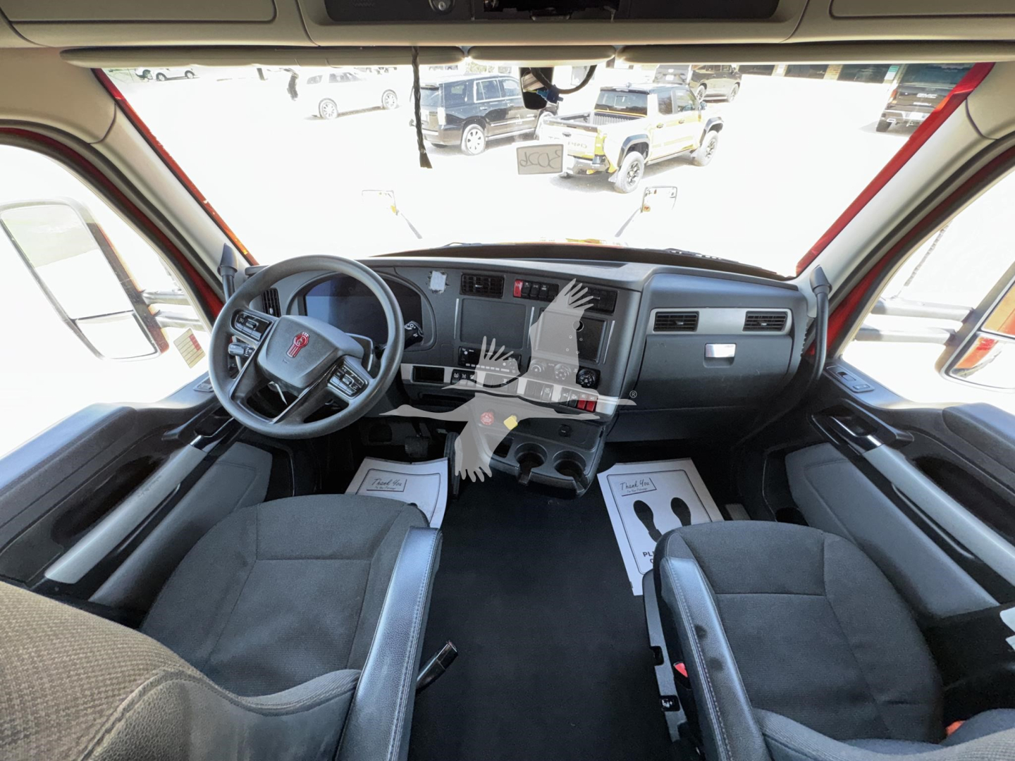 Driver's seat and dashboard view of a Kenworth truck cabin with steering wheel, center console, and front seats; no visible text readable.
