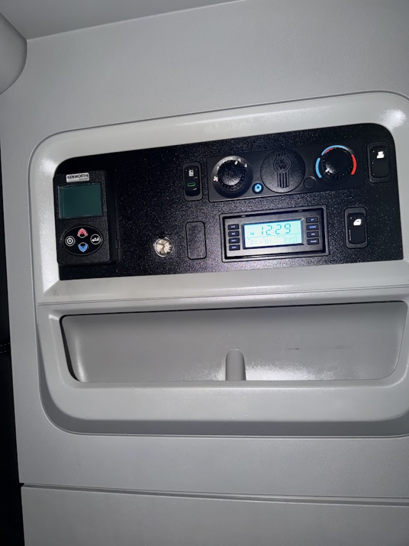 Truck cab dashboard showing control cluster with small display and circular knobs; air vent, switchgear, and digital readout visible.
