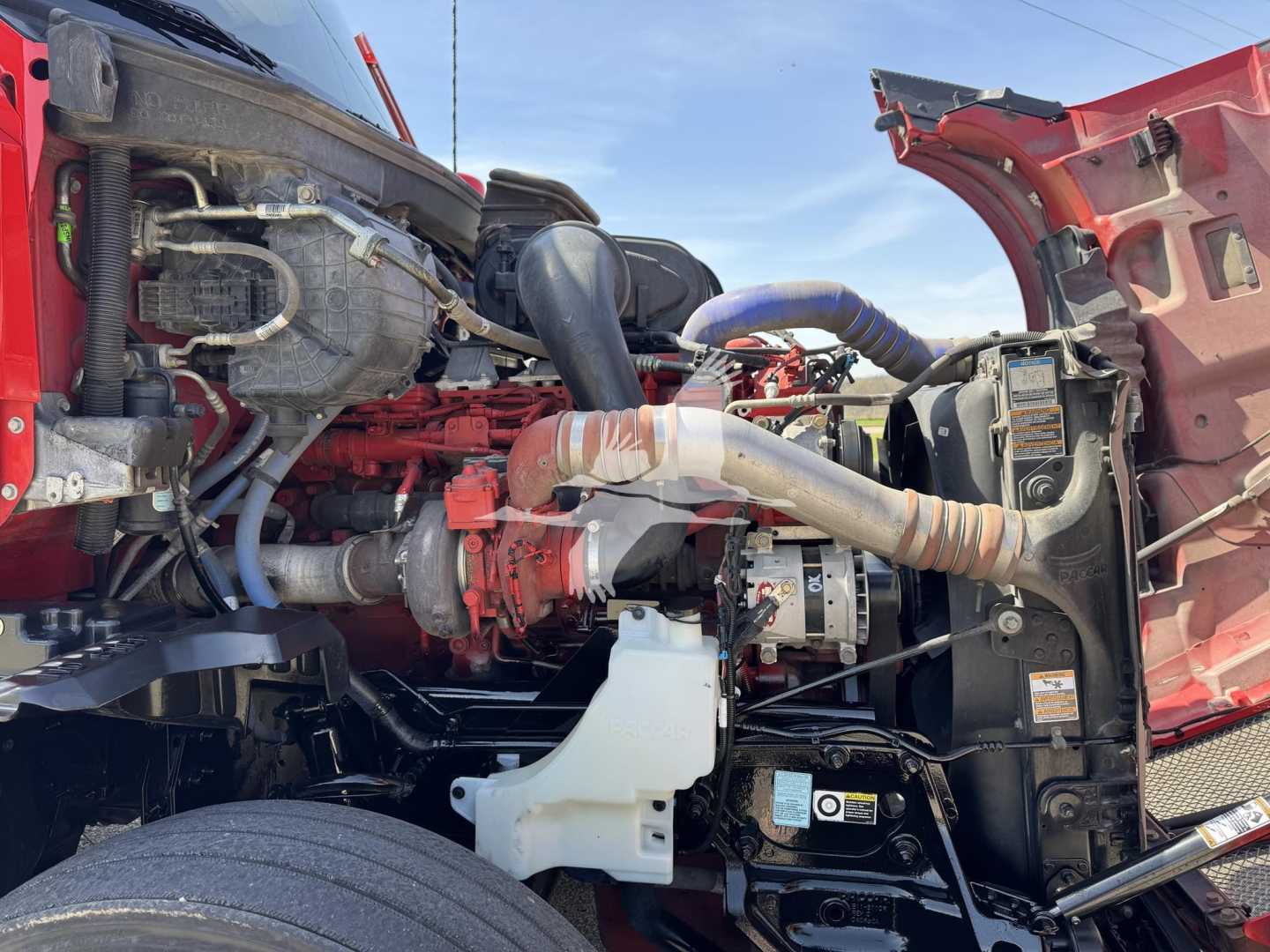 Kenworth T680 engine bay showing red engine block, intake tubing, hoses, and coolant reservoir; various components and wiring visible in open hood.