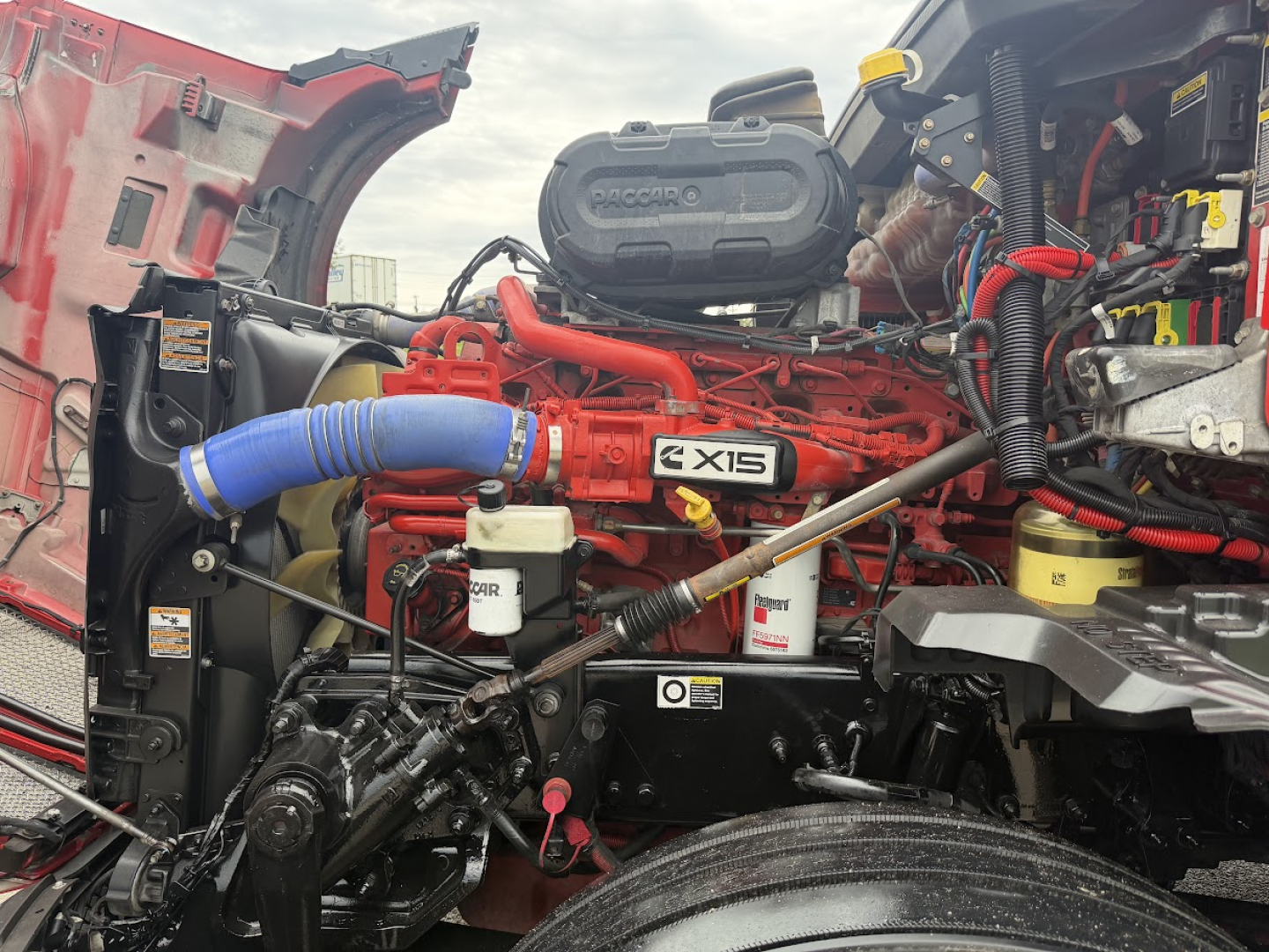 Kenworth T680 engine bay with red engine block labeled X15 and visible blue turbo hose, various hoses, caps, and wiring harnesses.