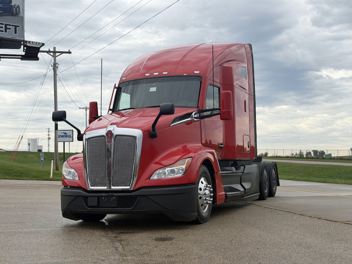 Red Kenworth T680 truck in a parking lot with a large chrome grille and dual mirrors; front-left view shows aggressive headlights.