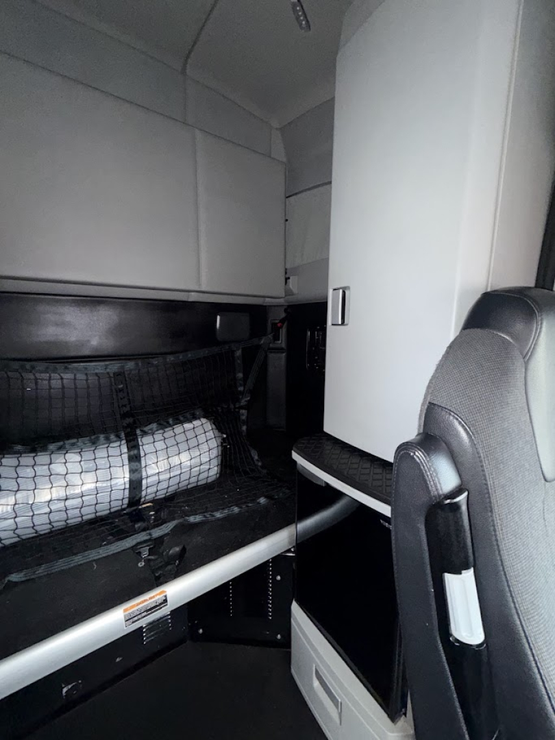 Kenworth T680 cab interior with sleeper area and storage under bunk; dashboard and seat visible with black and gray trim.