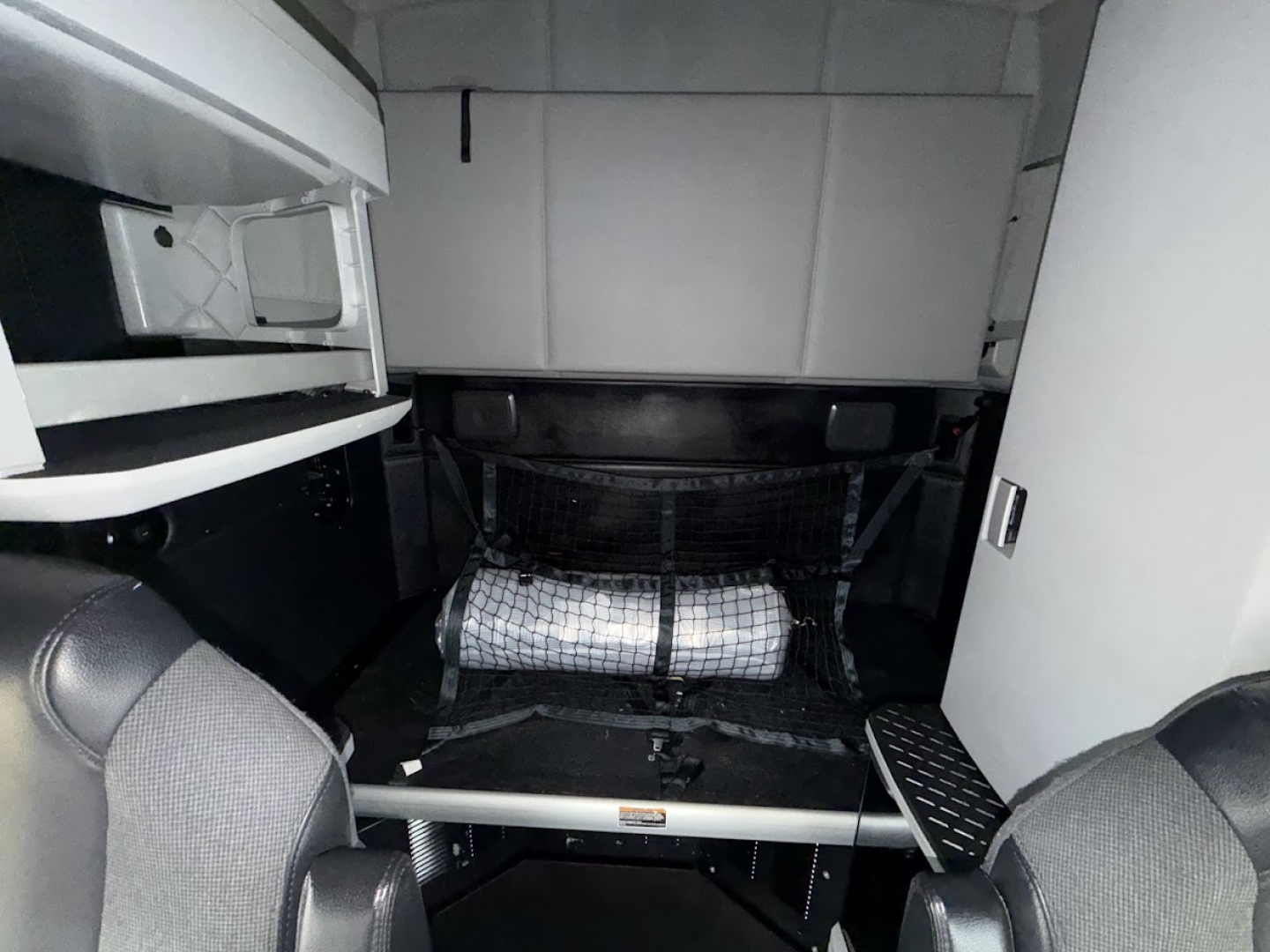 Cab area of a 2023 Kenworth T680 interior showcasing sleeper area with storage nets and steps, cockpit seating visible.