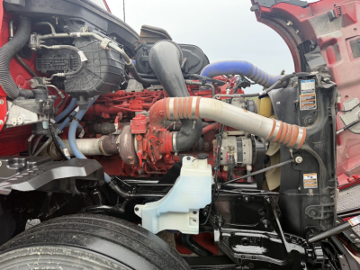 Kenworth T680 engine bay showing large red engine with turbocharger, blue hoses, radiator and coolant reservoir, white coolant overflow tank, and various mechanical components.