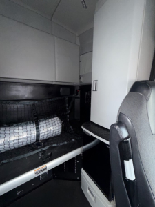 Kenworth T680 cab interior with sleeper area and storage under bunk; dashboard and seat visible with black and gray trim.