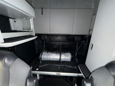 Cab area of a 2023 Kenworth T680 interior showcasing sleeper area with storage nets and steps, cockpit seating visible.