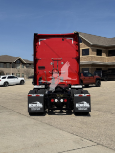 Kenworth T680 rear view showing red sleeper door, bumper mud flaps, and license plate area. VIN/stock not visible.