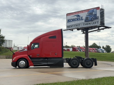 Kenworth T680 semi-truck in red with sleeper cab, side view, bumper and wheel set visible; billboard in background advertising Kordell with truck graphic.