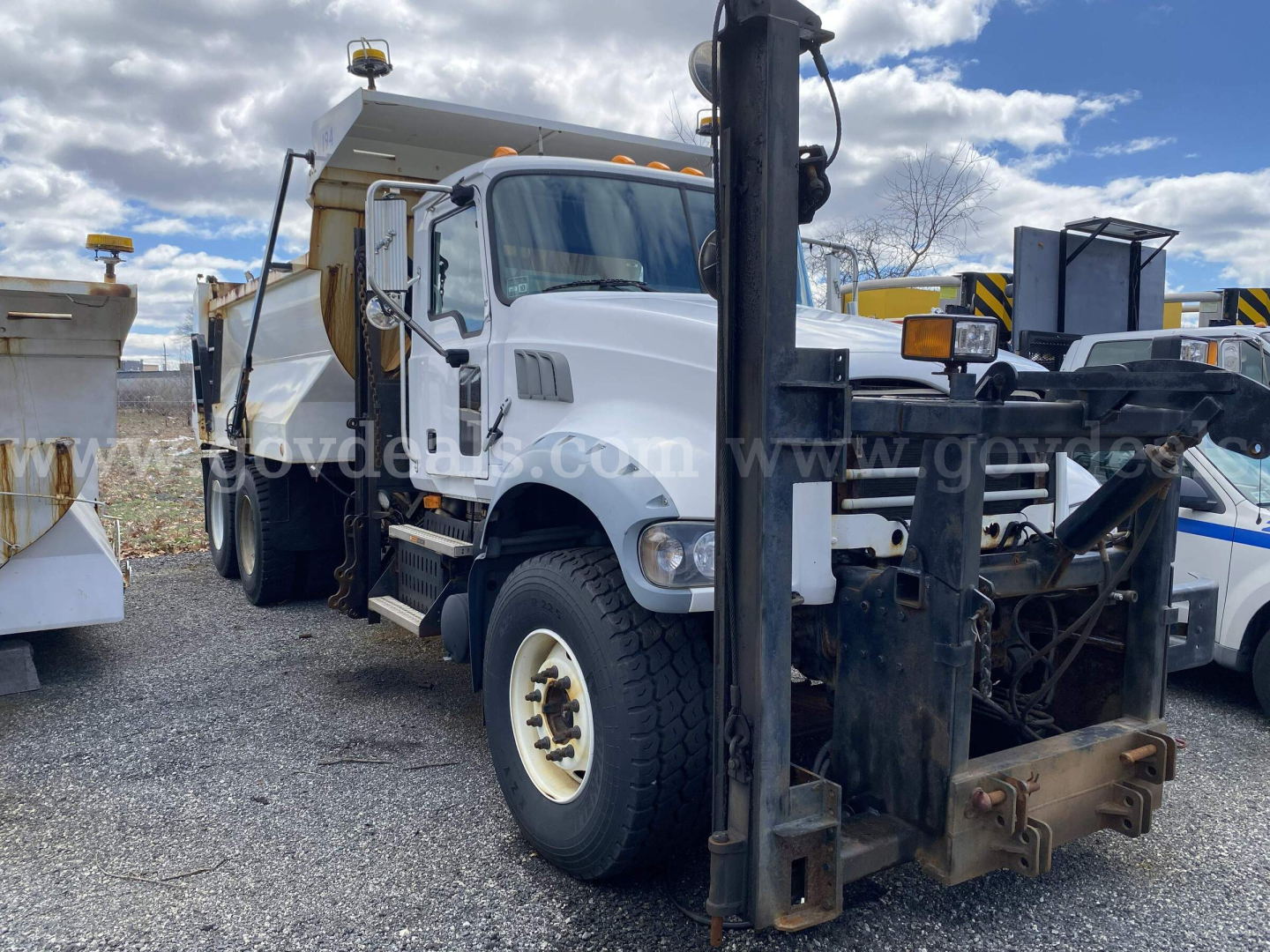 Dump truck front view showing Mack GU713 with attached front mount forks and hydraulic system. Visible text includes stock number and VIN partially on image watermark, and vehicle layout.