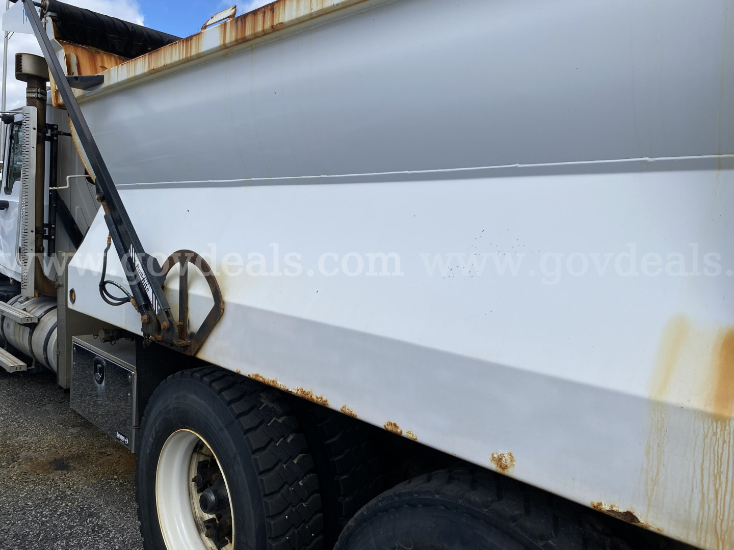 Dump truck side view showing rust spots along lower body and wheel with white rim, hydraulic mechanism and ladder area visible.