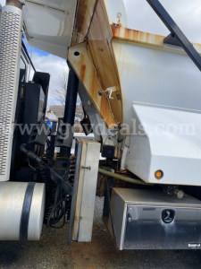 Mack GU713 dump truck rear side showing hydraulic hopper and rust-stained metal panels, VIN plate area not visible, with truck cab and fuel tank in view.