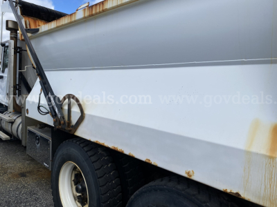 Dump truck side view showing rust spots along lower body and wheel with white rim, hydraulic mechanism and ladder area visible.