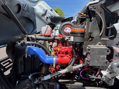 Mack MD6 engine bay showing red engine, blue coolant hose, various hoses, wiring harness, and diagnostic gauge cluster.