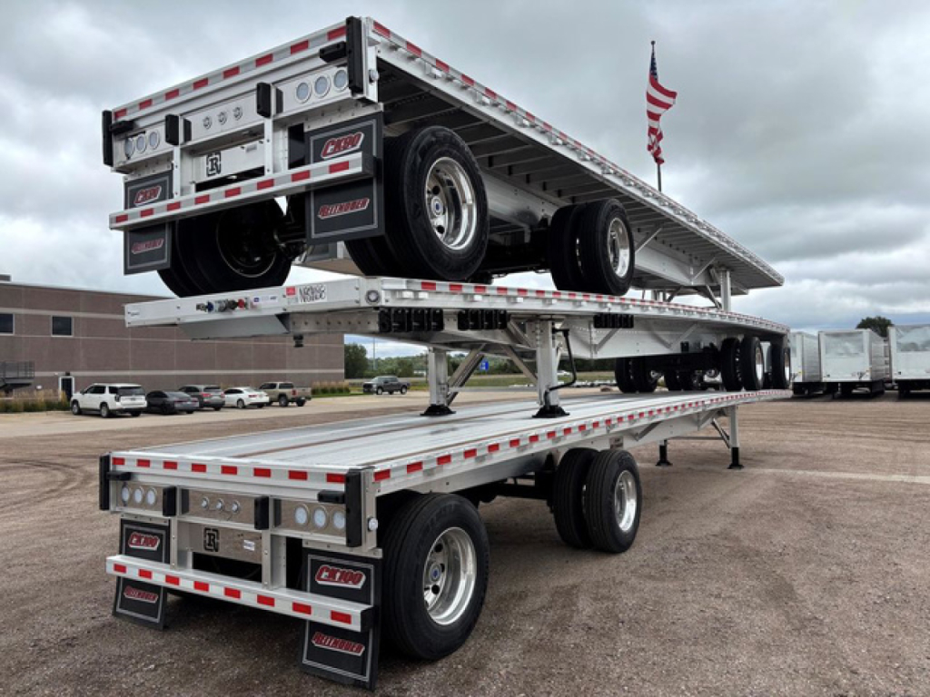 Flatbed trailer stacked on a two-tier hydraulic trailer with white frame and red/white reflective tape, dual rear axle, American flag in background.
