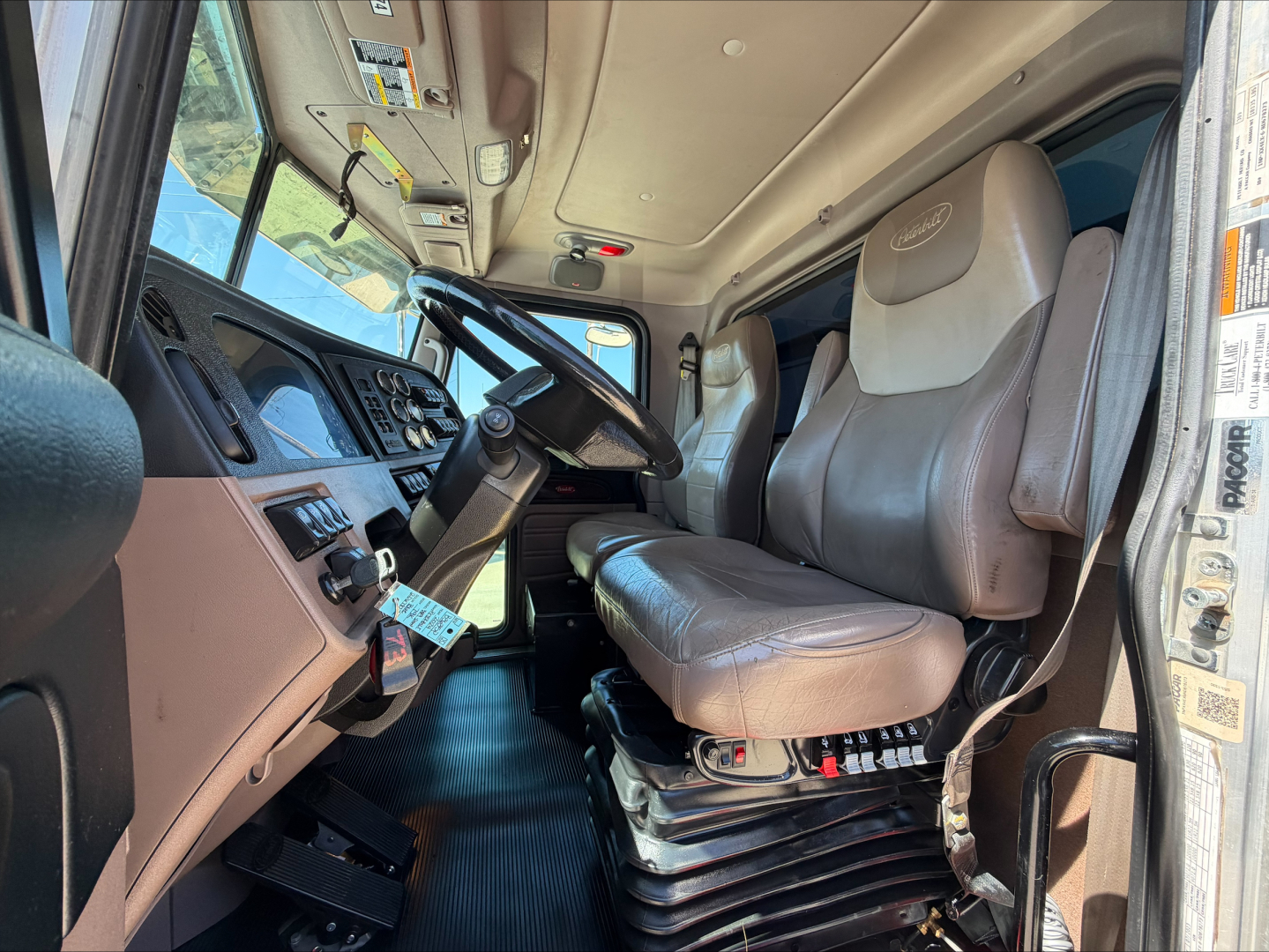 Truck cab interior of a 2024 Peterbilt 389 with tan leather seat, steering wheel, dashboard, and gear controls visible.