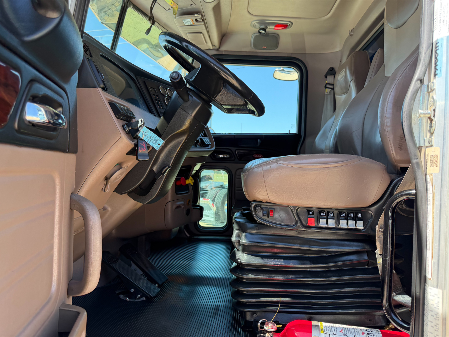 Truck cab interior showing steering wheel, driver's seat with multi-lever controls, dashboard, pedals, and sunlit windshield.