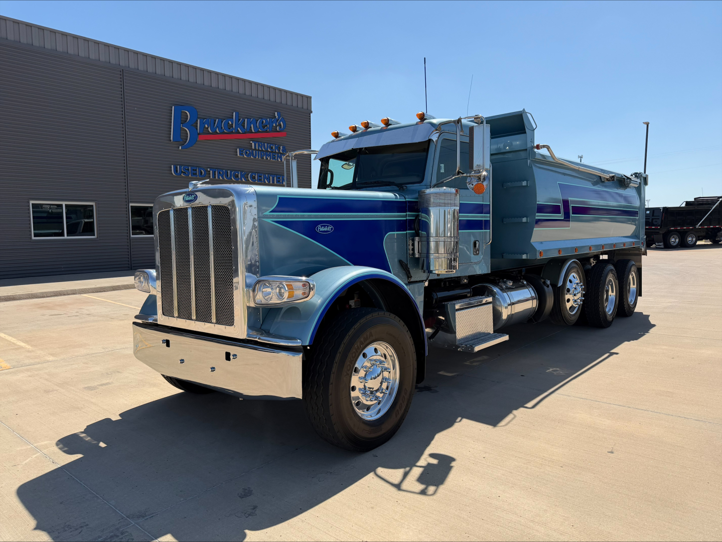 Peterbilt dump truck in blue and silver, front three-quarter view, chrome grille, dual exhaust, dump bed, daytime at dealership