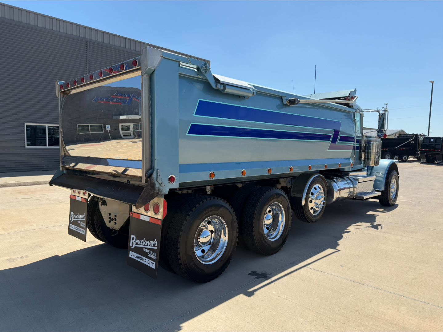 Peterbilt 389 dump truck rear and side view showing blue and silver dump bed with chrome wheels and dual rear axles.