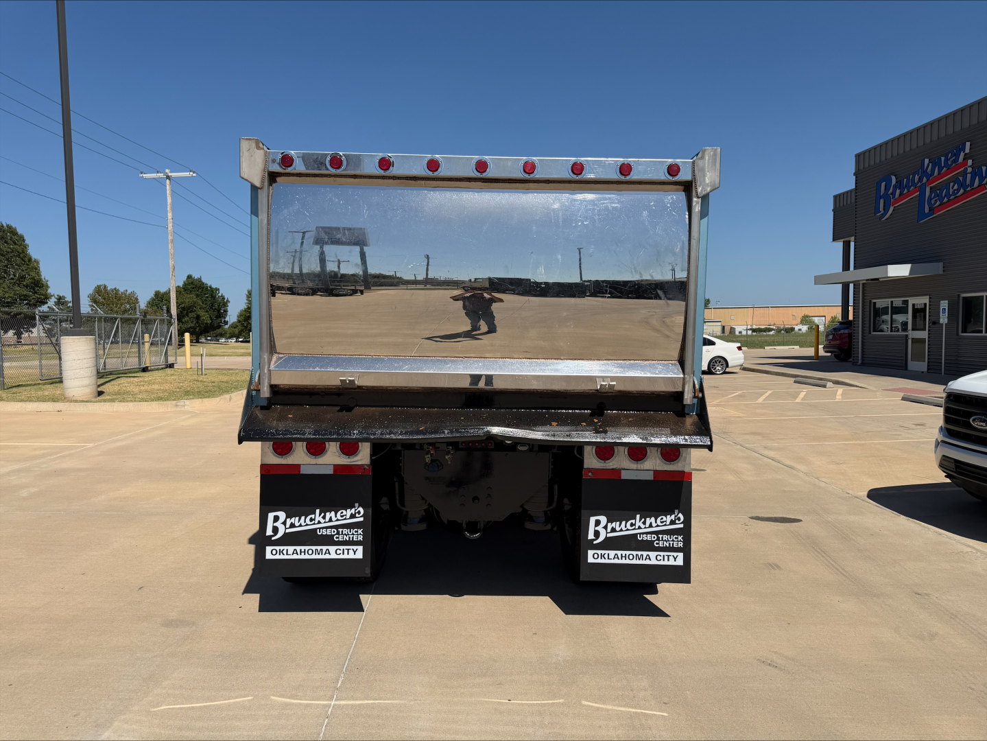 Rear view of a dump truck with reflective tailgate and dual mud flaps showing Bruckner's Oklahoma City dealership decals; wall of truck bed and sand transfer area in background.