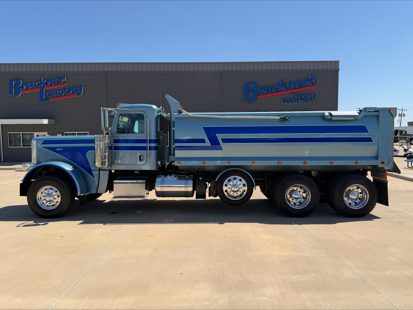 Peterbilt 389 dump truck side view showing blue and silver paint with chrome, fuel tanks, and dual rear axles.