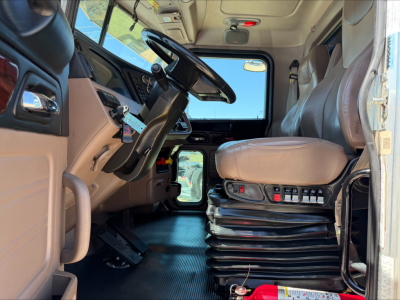 Truck cab interior showing steering wheel, driver's seat with multi-lever controls, dashboard, pedals, and sunlit windshield.