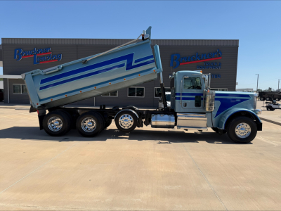 Peterbilt 389 dump truck in action with trailer, side view showing dump bed raised. Brand markings and paint scheme visible on cab and dump bed; background shows dealership building.