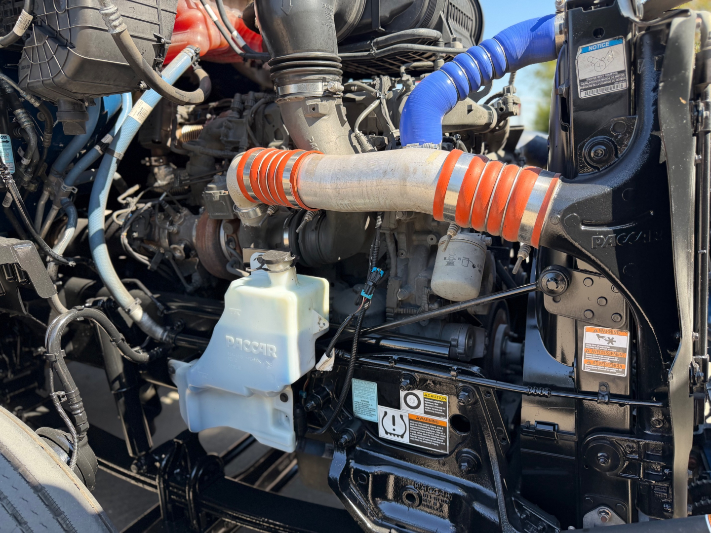 Kenworth T680 engine bay showing coolant reservoir, orange intake hose, blue hose, and black engine components.