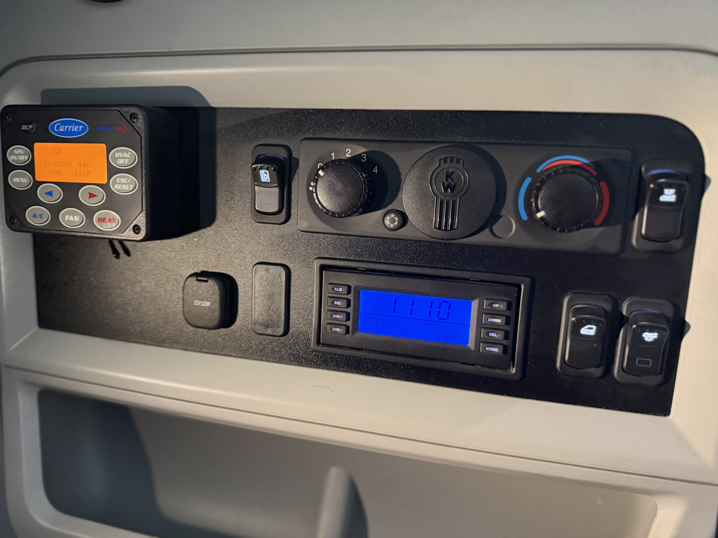 Kenworth T680 cab interior showing dashboard controls and HVAC knobs with blue digital display and Carrier device mounted on left.