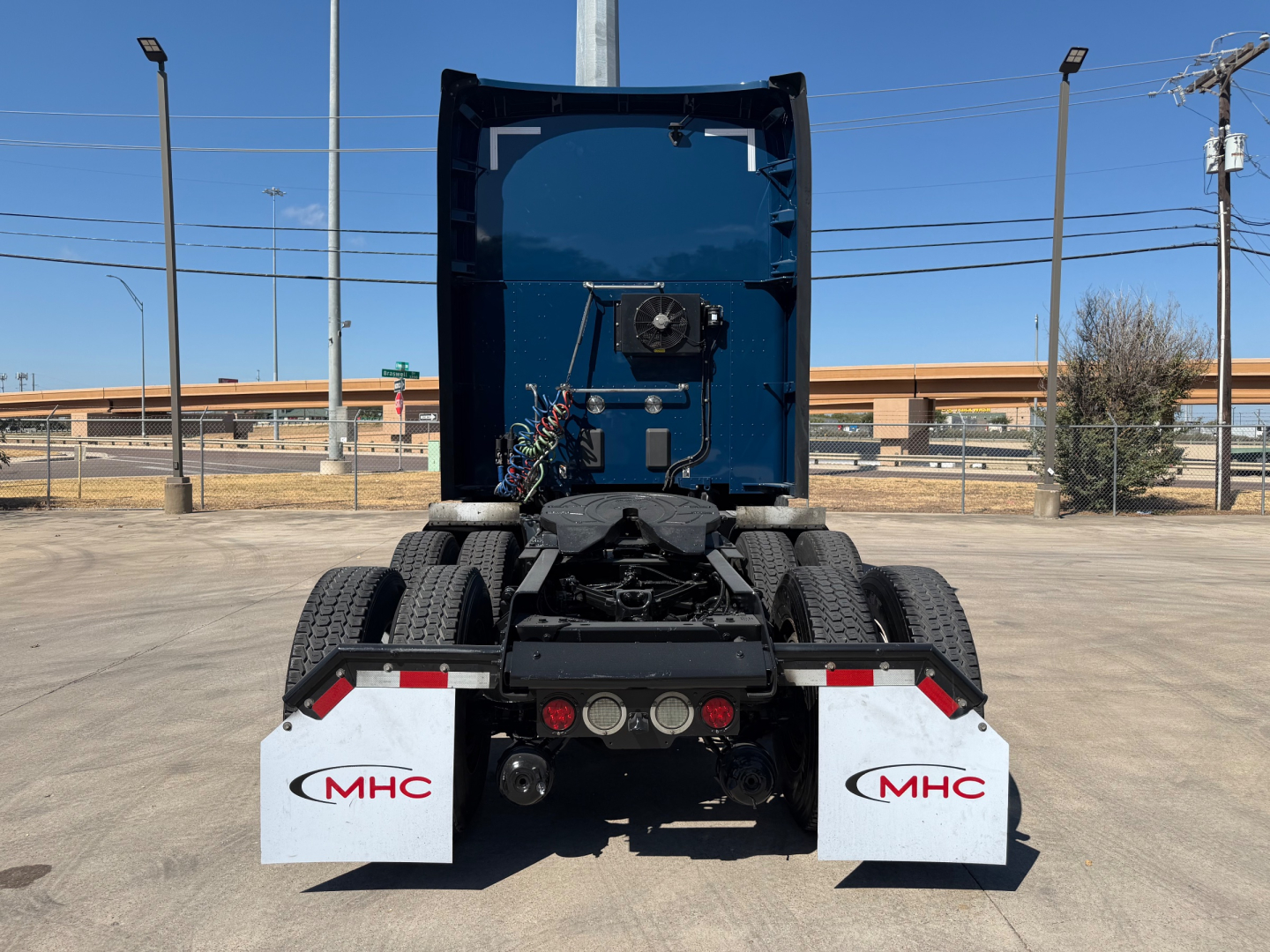 Rear view of a Kenworth truck tractor chassis showing mudflaps with MHC logos and rear lights.