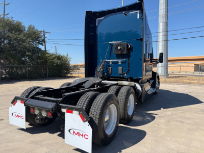 Kenworth T680 tractor rear view showing dual rear axles, fifth wheel, mud flaps with MHC branding, blue cab, and visible hydraulic lines and cooling unit on the back of the cab.