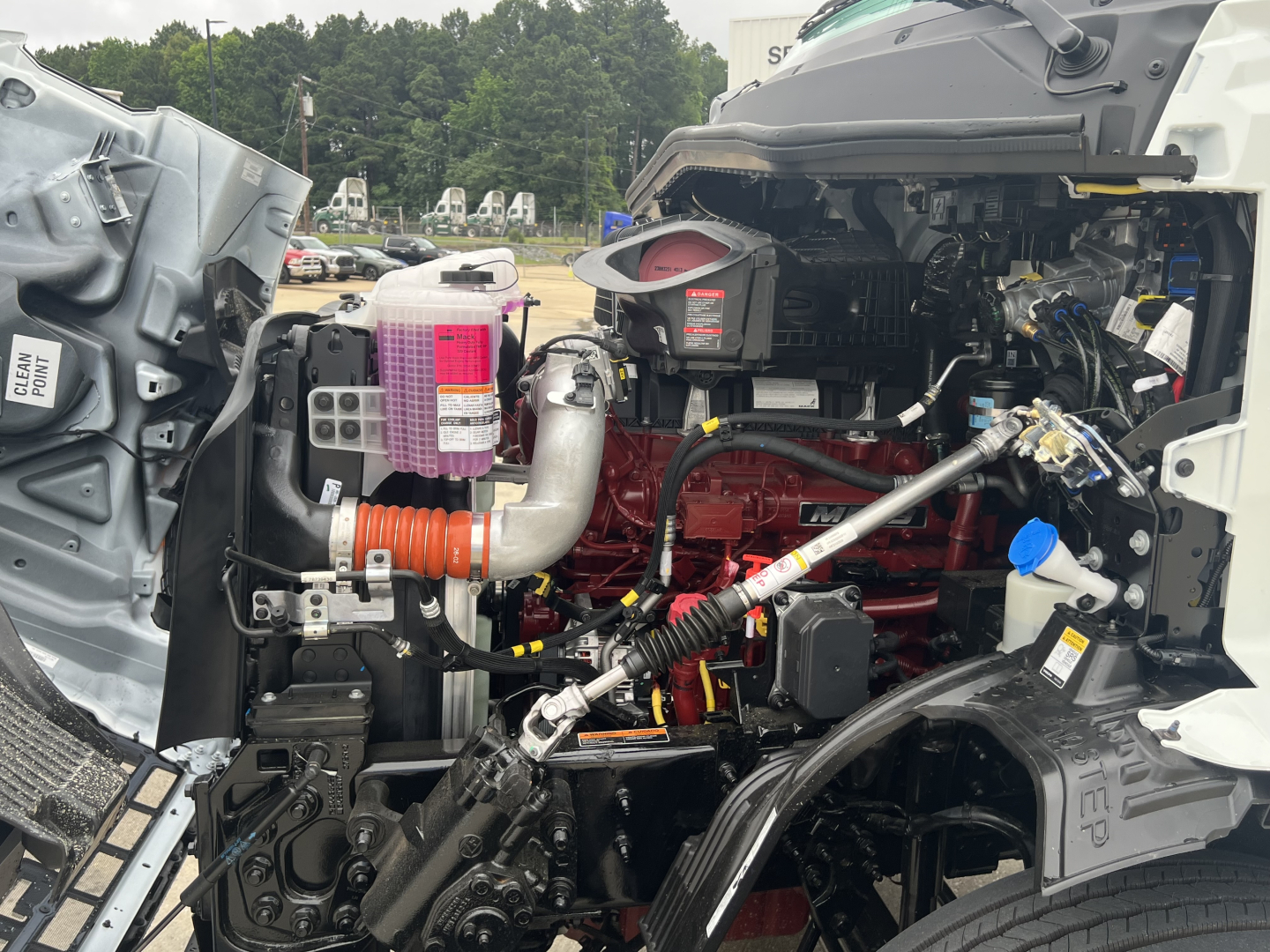 Front left engine bay of a 2027 Mack AN64T conventional daycab showing engine components, hoses, reservoirs, and wiring harnesses.