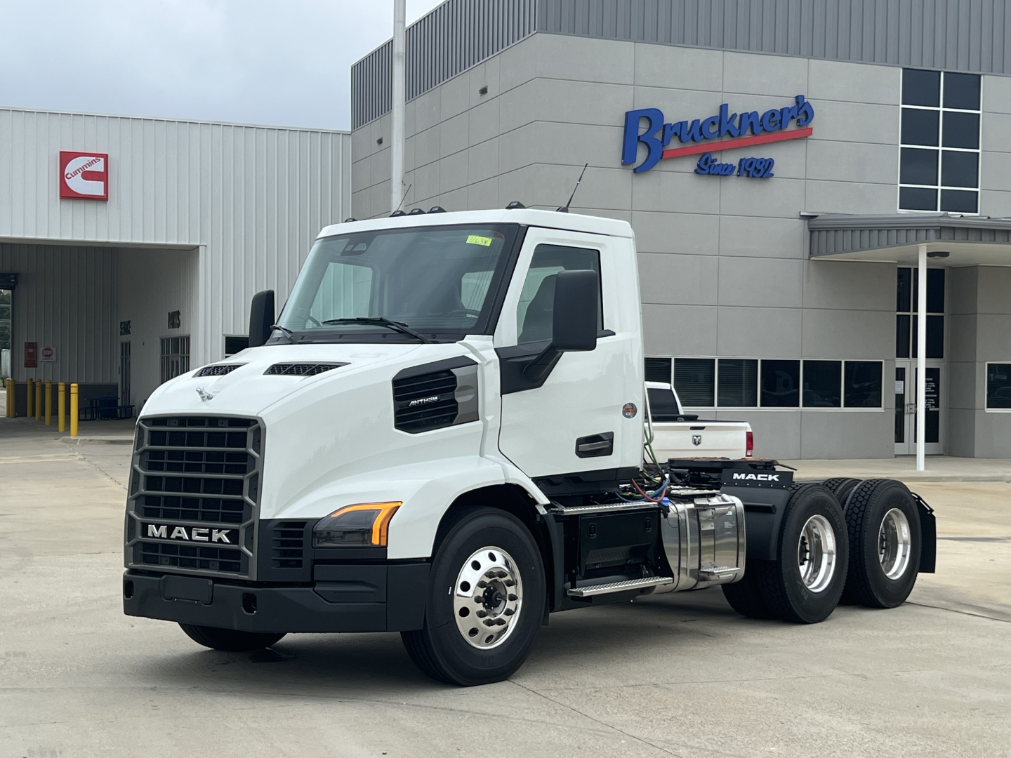 Mack AN64T conventional daycab truck viewed from front-left in a dealership lot, blank white cab, black grille, dual rear axles, fuel tanks, and Brackners dealership building in background.