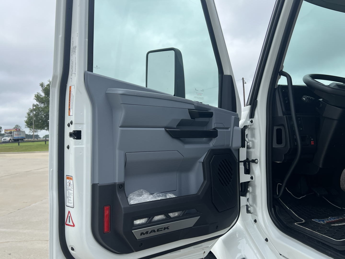 Truck cab interior door panel and open driver door showing Mack branding plate and control panels.