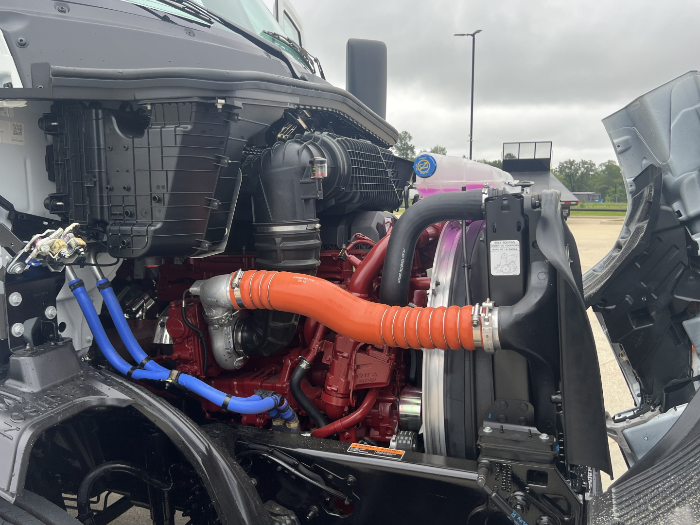 Truck engine bay of a 2027 Mack AN64T showing orange radiator hose, blue coolant lines, black radiator, and red engine components.