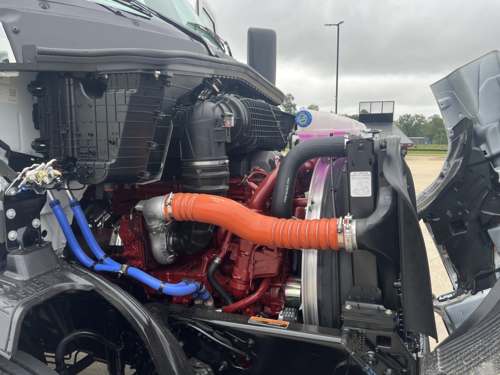 Truck engine bay of a 2027 Mack AN64T showing orange radiator hose, blue coolant lines, black radiator, and red engine components.
