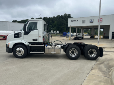 Truck chassis and cab of a 2027 Mack AN64T conventional daycab showing side profile, sensor cab, fuel tanks, and dual rear axles. Visible text on the image includes 'SERVICE' on the building.