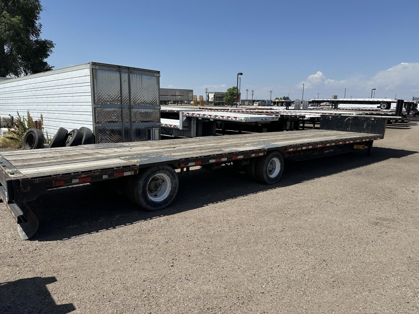 Trailer showing a long flatbed with dual rear axles and a freight trailer in background. Brand-new deck with wooden planks and metal frame. Clear sky.